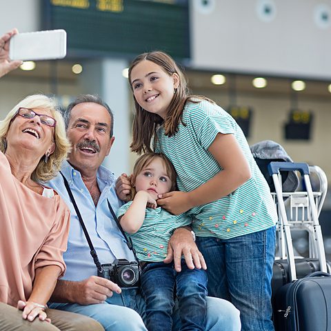 Grandparents and grandchildren taking a selfie at an airport departure lounge with suitcases, traveling to Italy