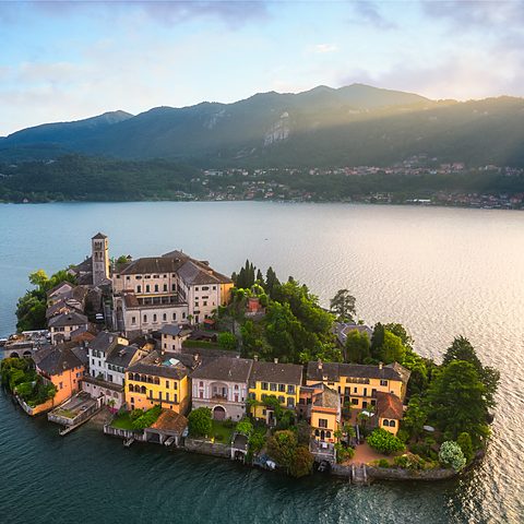 A picturesque island crowned by a bell tower rising from the calm waters of a Piedmont lake at dawn, Italy